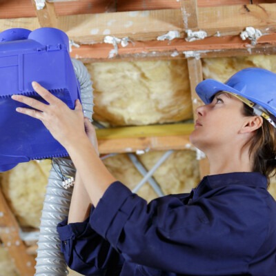 Technicienne Innovasun en train d’installer un caisson de VMC dans les combles d’une maison dans le var, portant un casque de protection et vérifiant le raccordement des gaines de ventilation.